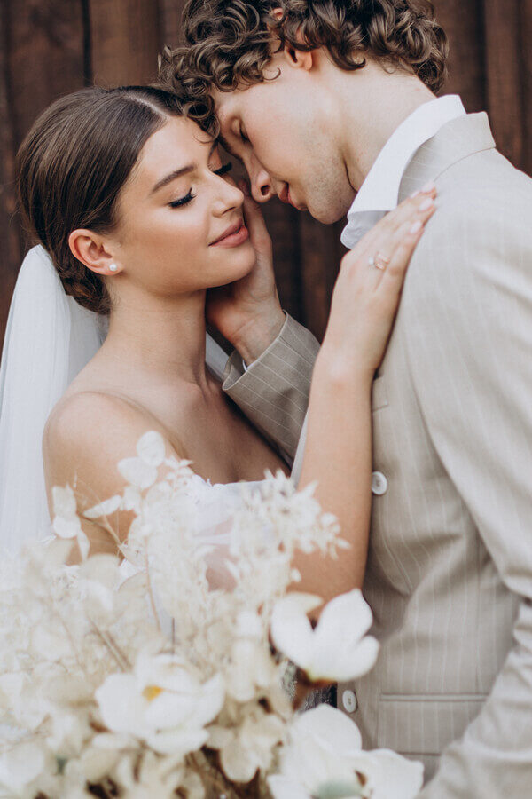 Los novios frente a frente junto a una pared de madera; ella lleva velo y vestido palabra de honor, y apoya la mano en el hombro de él