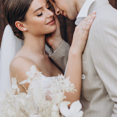 Los novios frente a frente junto a una pared de madera; ella lleva velo y vestido palabra de honor, y apoya la mano en el hombro de él