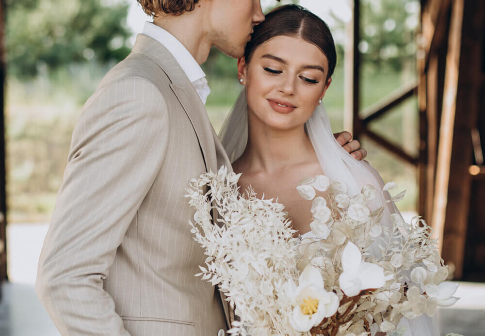 El novio besa a la novia en la sien mientras ella sostiene un ramo de flores secas blancas
