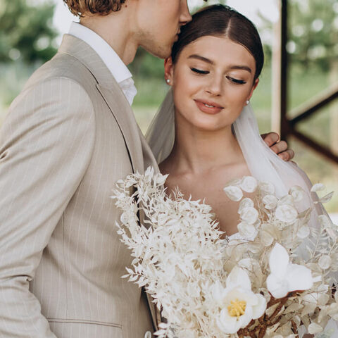 El novio besa a la novia en la sien mientras ella sostiene un ramo de flores secas blancas