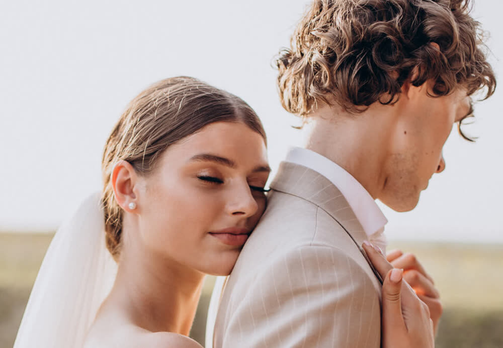 La novia apoya la mejilla en el hombro del novio, de espaldas al campo y bajo un cielo despejado