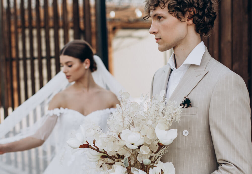 El novio de perfil con el ramo en la mano; detrás, la novia camina con el velo al viento