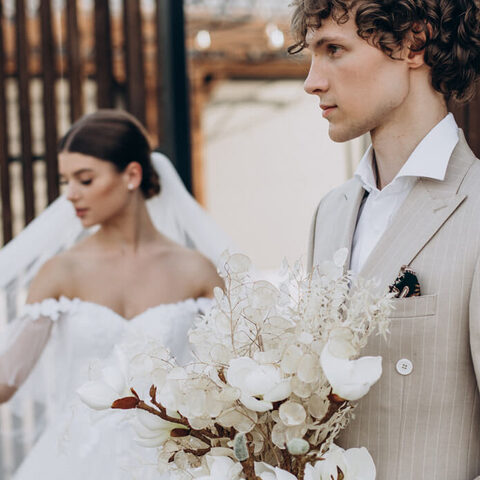 El novio de perfil con el ramo en la mano; detrás, la novia camina con el velo al viento